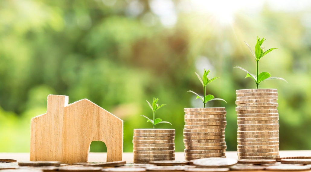 Little stacks of coins with plants at the top next to a wooden plate in the form of a house